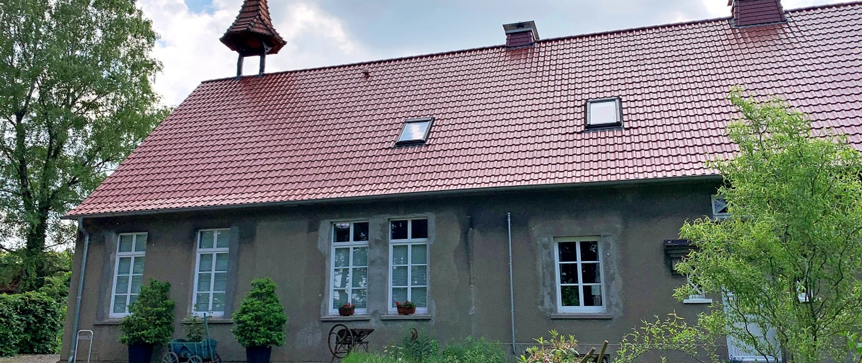 Altes Schulgebäude mit rotem Ziegeldach, Glockenturm und gepflegtem Garten, von Bäumen umgeben.