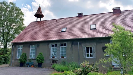 Alte Volksschule Rehmerloh Altes Schulgebäude mit rotem Ziegeldach, Glockenturm und gepflegtem Garten, von Bäumen umgeben.