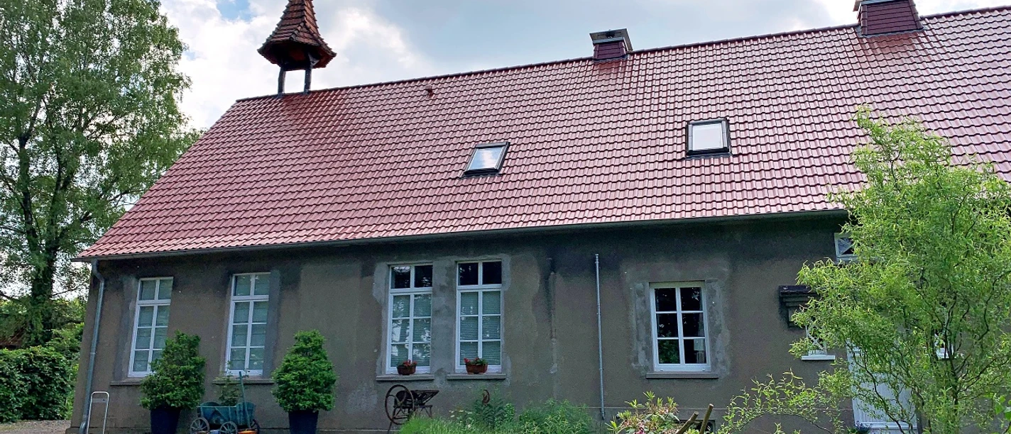 Alte Volksschule Rehmerloh Altes Schulgebäude mit rotem Ziegeldach, Glockenturm und gepflegtem Garten, von Bäumen umgeben.