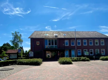 Rathaus Wiefelstede.jpg Das Rathaus Wiefelstede zeigt ein einstöckiges Ziegelgebäude mit großen Fenstern und einem Schieferdach. Ein gepflasterter Vorplatz führt zum Eingang.