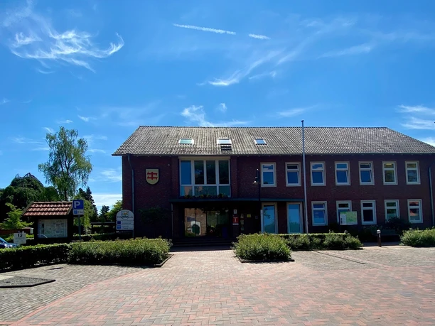 Rathaus Wiefelstede.jpg Das Rathaus Wiefelstede zeigt ein einstöckiges Ziegelgebäude mit großen Fenstern und einem Schieferdach. Ein gepflasterter Vorplatz führt zum Eingang.