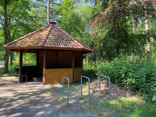 Rastplatz Mansholter Holz.jpg Ein kleiner Rastplatz mit einem hölzernen Pavillon und roten Ziegeln, umgeben von dichtem Grün.