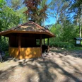 Rastplatz Mansholter Holz2.jpg Ein hölzerner Pavillon mit schattenspendendem Dach steht an einem Waldweg, umgeben von grünen Bäumen und einem Fahrradständer.