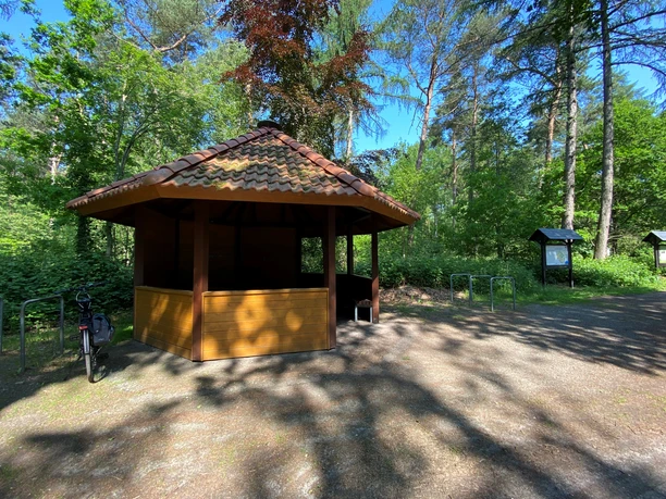 Rastplatz Mansholter Holz2.jpg Ein hölzerner Pavillon mit schattenspendendem Dach steht an einem Waldweg, umgeben von grünen Bäumen und einem Fahrradständer.