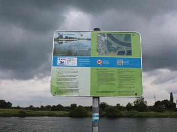 Ein Informationsschild in ländlicher Flusslandschaft unter bewölktem Himmel, nahe dem Wasser positioniert.