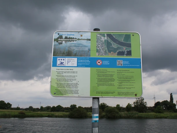 Ein Informationsschild in ländlicher Flusslandschaft unter bewölktem Himmel, nahe dem Wasser positioniert.