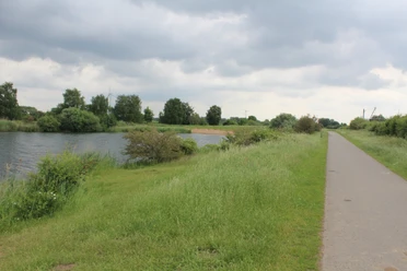 Flussufer mit Fuß- und Radweg, umgeben von saftigem Grün; im Hintergrund Windmühlen unter bewölktem Himmel.