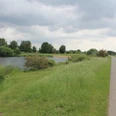 Flussufer mit Fuß- und Radweg, umgeben von saftigem Grün; im Hintergrund Windmühlen unter bewölktem Himmel.