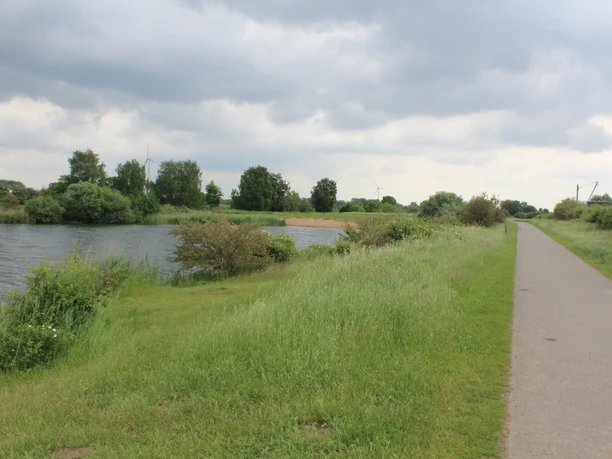 Flussufer mit Fuß- und Radweg, umgeben von saftigem Grün; im Hintergrund Windmühlen unter bewölktem Himmel.