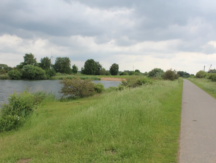 Flussufer mit Fuß- und Radweg, umgeben von saftigem Grün; im Hintergrund Windmühlen unter bewölktem Himmel.