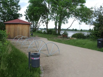 Ein kleiner Holzpavillon neben einem Flussuferweg, umgeben von grüner Vegetation und Fahrradständern.