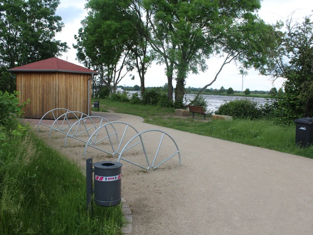 A small wooden pavilion next to a riverside path, surrounded by greenery and bike racks.