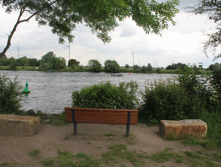 Eine Holzbank mit zwei Steinen am Weserrand, umgeben von grüner Vegetation und Blick auf Windräder.