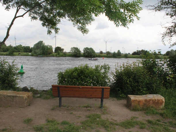 Reisegarten Weye Eine Holzbank mit zwei Steinen am Weserrand, umgeben von grüner Vegetation und Blick auf Windräder.