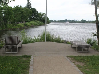 Holzbank am ruhigen Flussufer mit üppigem Grün und sanftem Wasserlauf unter bewölktem Himmel.