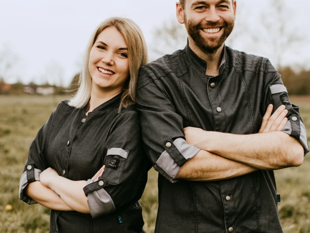BBQ Team Ammerland Das Bild zeigt ein lächelndes Paar im Freien, beide mit gekreuzten Armen und in Uniformen des BBQ Teams Ammerland.