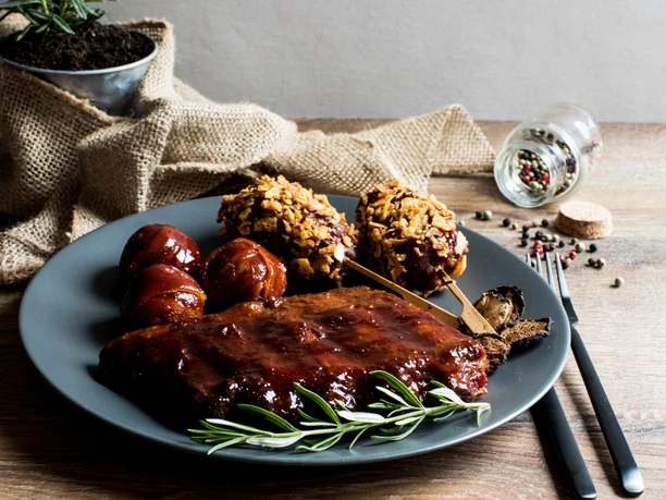 Grillvergnügen Ein grauer Teller mit saftigen Rippchen in Barbecuesoße, Kartoffelspießen und Kräutergarnitur.