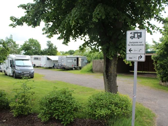 Wohnmobilstellplatz im Grünen mit mehreren geparkten Fahrzeugen und einem Hinweisschild am Wegesrand.