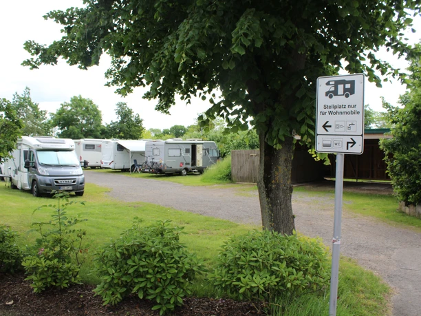 Wohnmobilstellplatz im Grünen mit mehreren geparkten Fahrzeugen und einem Hinweisschild am Wegesrand.