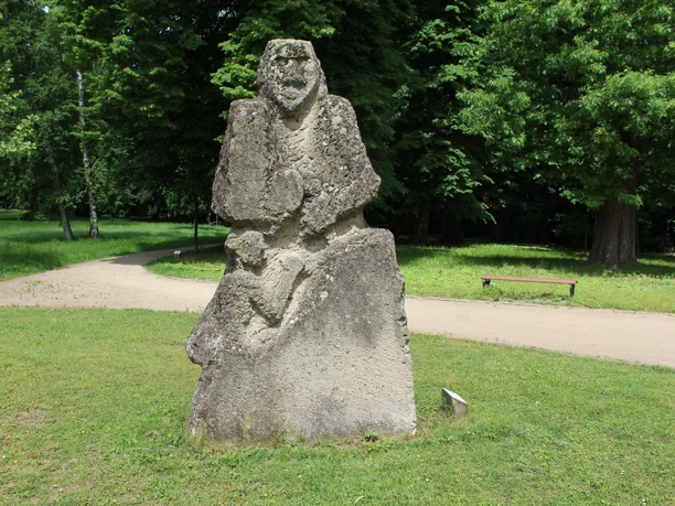 The stone sculpture "Kopfgeburt" stands on a lawn in Etelsen Park, surrounded by lush greenery.