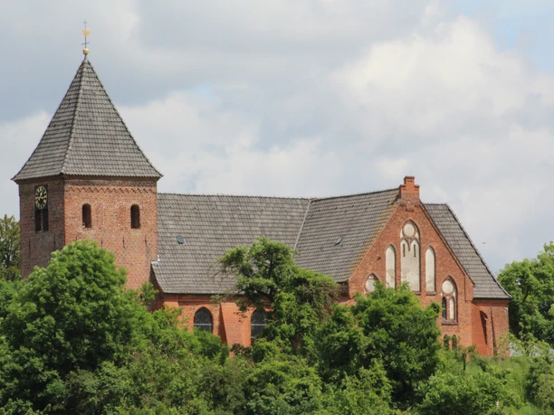 Backsteinkirche mit Glockenturm und Satteldach, umgeben von grünen Bäumen unter blauem Himmel.