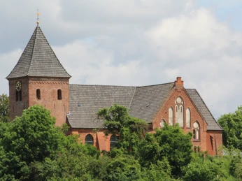Kirche Daverden Backsteinkirche mit Glockenturm und Satteldach, umgeben von grünen Bäumen unter blauem Himmel.