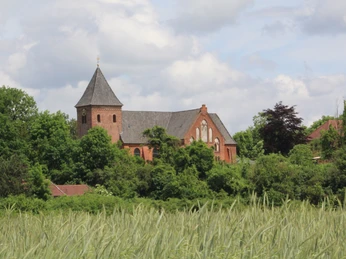 Rote Backsteinkirche mit Turm, umgeben von Bäumen; im Vordergrund weite Felder unter blauem Himmel.