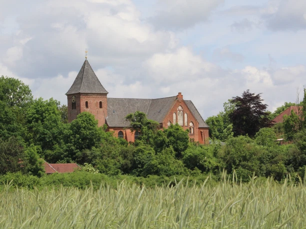 Rote Backsteinkirche mit Turm, umgeben von Bäumen; im Vordergrund weite Felder unter blauem Himmel.
