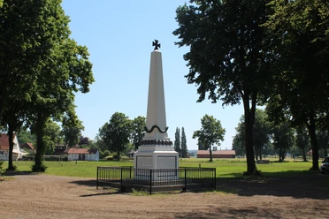Denkmal auf einem Marktplatz, umrahmt von Bäumen, mit einem schwarzen Zaun davor. Wiese im Hintergrund.