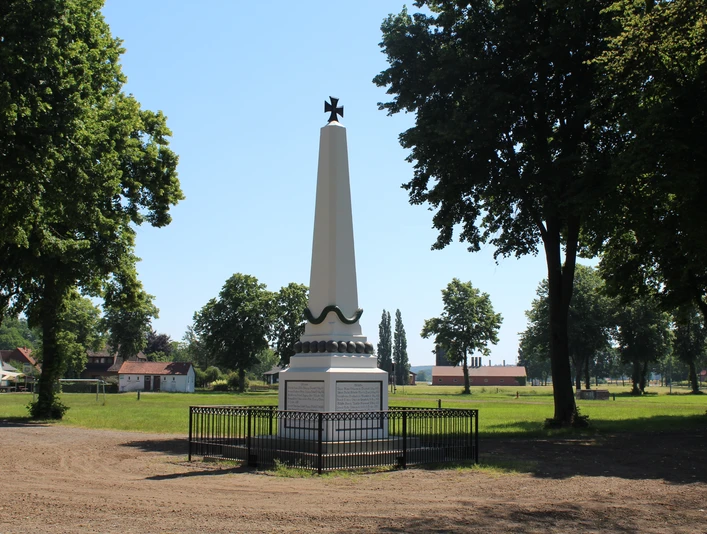 Denkmal Marktplatz Bruchhausen-Vilsen Denkmal auf einem Marktplatz, umrahmt von Bäumen, mit einem schwarzen Zaun davor. Wiese im Hintergrund.