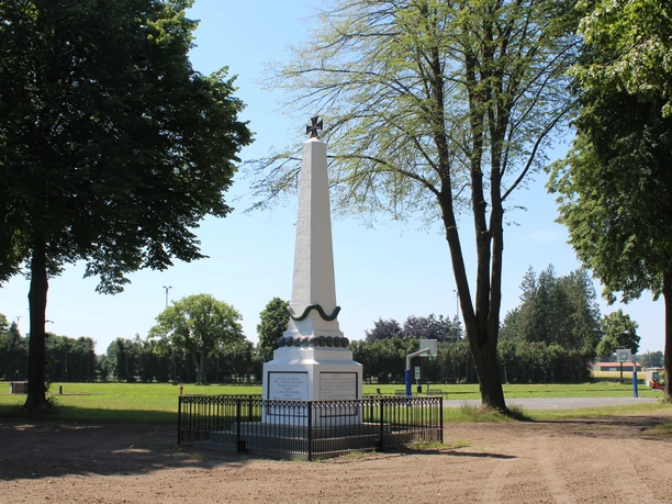 Ein weißer Obelisk mit Skulptur auf grünem Platz, umgeben von Bäumen, in Bruchhausen-Vilsen.