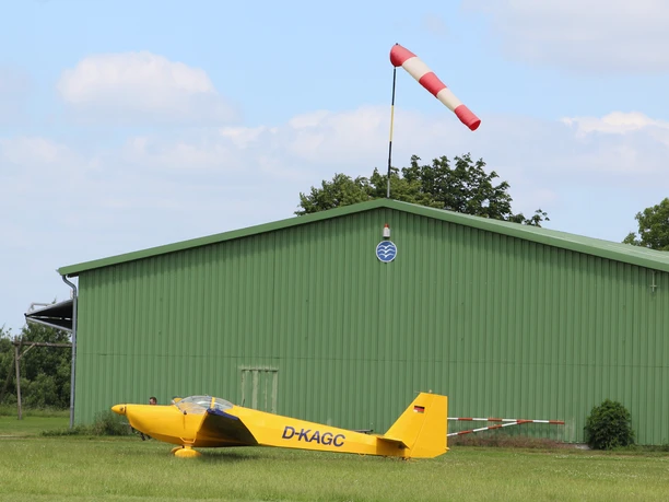 Gelber Segelflieger vor einer grünen Halle auf einem Flugplatz mit einer rot-weißen Windsack.