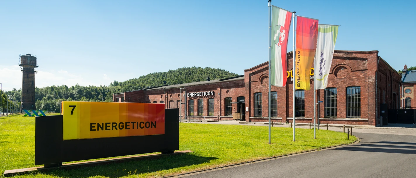 Eingangsbereich zu einem industriell aussehenden Gebäude mit Fahnen und einer Wiese davor.