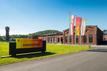 ENERGETICON Haupteingang Eingangsbereich zu einem industriell aussehenden Gebäude mit Fahnen und einer Wiese davor.