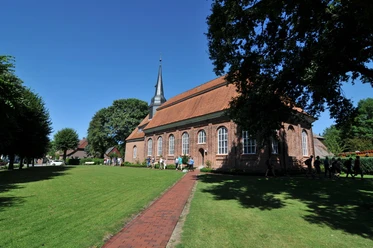Historische rote Backsteinkirche Emmaus in Neuhaus, umgeben von grünen Wiesen und blauem Himmel.
