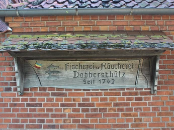 Holzschild an einer roten Backsteinwand mit der Inschrift „Fischerei u. Räucherei Dobberschütz“.