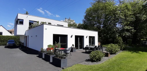 20210530_114330_Gebäudeansicht.jpg Modernes, weißes Einfamilienhaus mit Flachdach, großen Fenstern und Terrasse in grüner Umgebung.