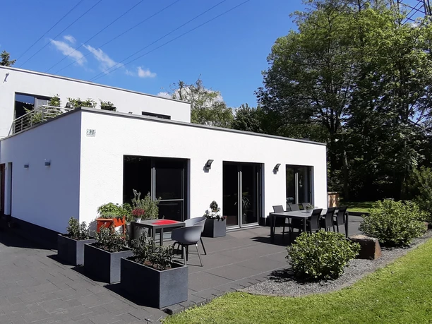 20210530_114330_Gebäudeansicht.jpg Modernes, weißes Einfamilienhaus mit Flachdach, großen Fenstern und Terrasse in grüner Umgebung.