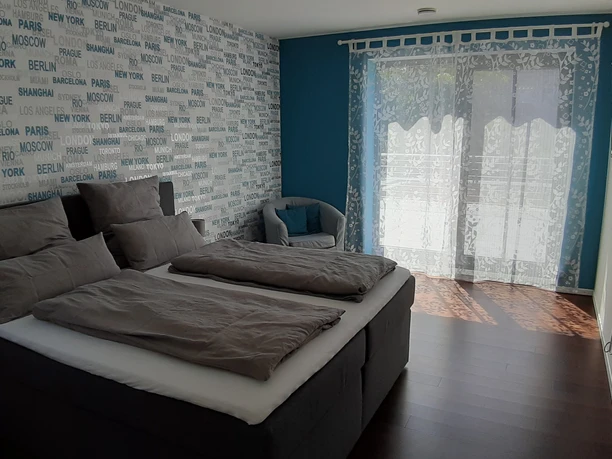 20210530_113616_Schlafzimmer.jpg Helles Schlafzimmer mit Doppelbett, grauer Bettwäsche und Texttapete, Fenster mit weißen Vorhängen.