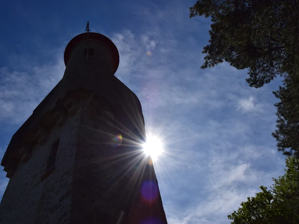 Der Aussichtsturm im Gegenlicht <img alt="Steinerner Turm im Gegenlicht, Sonne scheint, Bäume daneben, blauer Himmel mit Wolken." />