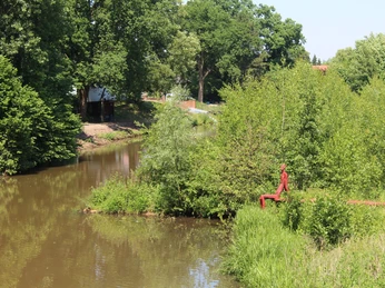 Eine rote Skulptur eines sitzenden Mannes am Ufer eines Flusses, umgeben von üppigen Bäumen.