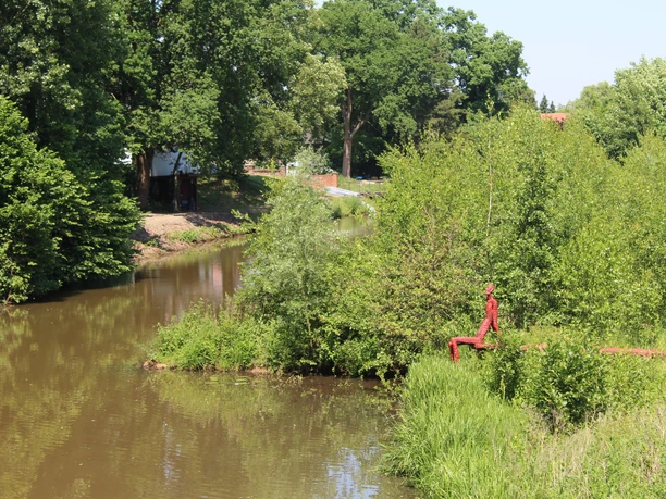 Eine rote Skulptur eines sitzenden Mannes am Ufer eines Flusses, umgeben von üppigen Bäumen.