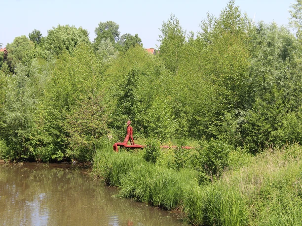 Eine rote Skulptur steht am Flussufer, umgeben von üppigem Grün, inmitten einer friedvollen Landschaft.