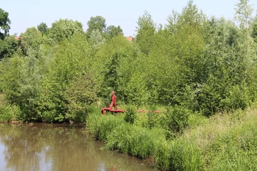 Eine rote Skulptur steht am Flussufer, umgeben von üppigem Grün, inmitten einer friedvollen Landschaft.