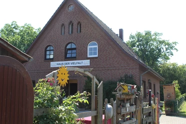 Haus der Vielfalt Backsteinhaus mit drei Fensterbögen und "Haus der Vielfalt"-Schild, umgeben von blühenden Pflanzen.