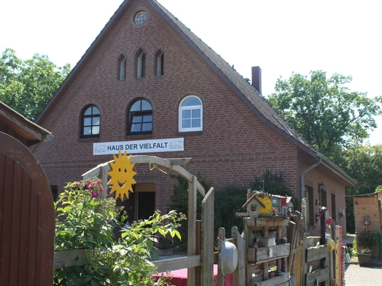 Haus der Vielfalt Backsteinhaus mit drei Fensterbögen und "Haus der Vielfalt"-Schild, umgeben von blühenden Pflanzen.
