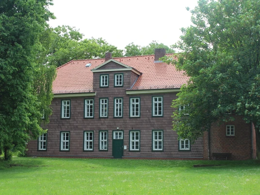 Historisches Gebäude aus rotem Backstein mit grünem Rasen, umrahmt von üppigen Bäumen.