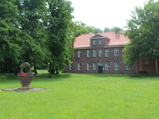 Historisches Backsteingebäude mit rotem Dach, umgeben von grüner Rasenfläche und schattenspendenden Bäumen.