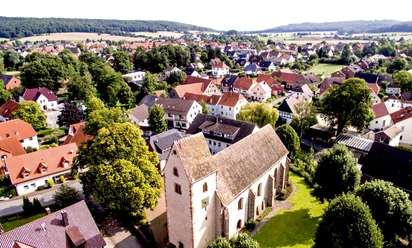 Langenholzhausen mit grünem Hügelpanorama, Kirche im Zentrum, umgeben von Wohnhäusern und Bäumen.