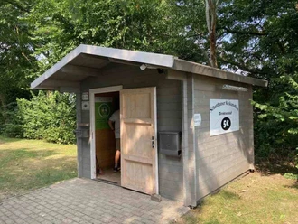 Hofladen Schatthaus Pewsum Graues Holzhäuschen unter Bäumen mit offener Tür. Ein Schild bewirbt Kartoffel-Direktverkauf.